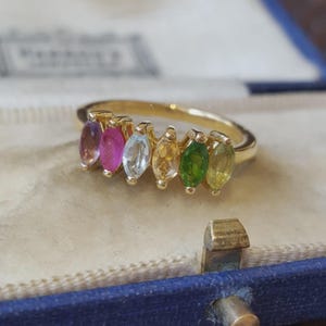 Può includere: Un anello in oro con sei pietre preziose taglio marquise disposte in fila. Le pietre presentano uno spettro di colori che include viola, rosa, azzurro, giallo, verde e oliva, incastonate in una semplice fascia in oro lucido.