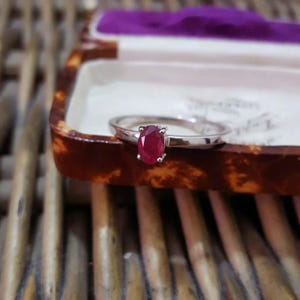 May include: A silver ring featuring an oval ruby gemstone. The ring has a sleek, polished band. The ruby is a rich red hue, and the ring is presented in a vintage jewellery box.