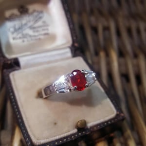 Może przedstawiać: Srebrny pierścionek z czerwonym owalnym kamieniem szlachetnym otoczonym mniejszymi przezroczystymi kamieniami. Pierścionek znajduje się w małym pudełku.