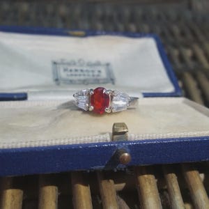 May include: A silver ring featuring a central oval red gemstone, with two rectangular clear stones either side. The ring is presented in a blue jewellery box with a white interior.