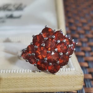 May include: A silver-toned ring featuring a cluster of numerous round, deep orange-red faceted stones arranged in a dense, multi-layered formation. Each stone is secured by small metal prongs, creating a textured, sparkling surface across the top.