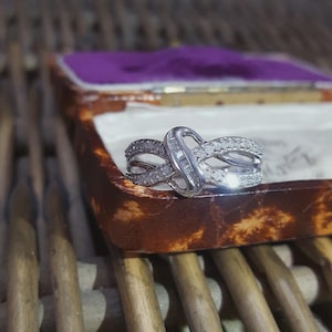 Op de afbeelding: Een zilveren ring met een oneindig knoopontwerp, bezet met kleine, heldere edelstenen. De ring wordt gepresenteerd in een vintage bruine sieradendoos met een paars interieur. De doos staat op een houten oppervlak.