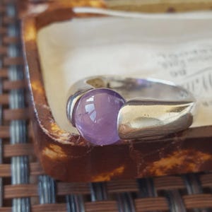 May include: A silver ring featuring a smooth, round, light purple gemstone set between two polished, curved metal ends. The band has a wide, solid construction that tapers toward the back, showcasing a reflective, metallic finish.