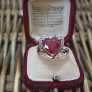 May include: A silver ring featuring a heart-shaped, deep red gemstone, encircled by a halo of small, clear stones. The ring is presented in an open, vintage red velvet jewellery box. The box's interior is cream-coloured.