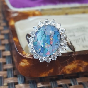 May include: A silver-toned ring featuring a large, oval-shaped centerpiece composed of inlaid blue and iridescent mosaic opal fragments, surrounded by a halo of small, clear, round-cut white stones set in a scalloped metal frame.