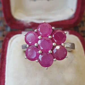 May include: A silver-toned ring featuring a cluster of seven round, faceted, vibrant pink stones arranged in a floral pattern. The stones are set in a metal prong setting, creating a circular cluster design that sits atop a smooth, polished metal band.