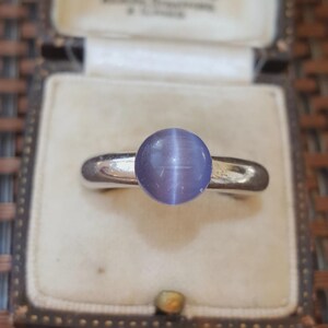 May include: A silver ring featuring a round, cabochon-cut, light purple cat's eye stone. The stone displays a distinct vertical chatoyant band of light across its centre. The ring band is smooth, polished, and metallic, showcasing a simple design.
