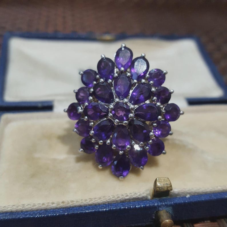 K&ouml;nnte beinhalten: Ein Silberring mit einem Cluster aus tiefvioletten Amethyst-Edelsteinen. Die Edelsteine sind tropfenf&ouml;rmig und in einem floralen Muster angeordnet. Der Ring wird in einer blauen Schmuckschatulle mit hellem beigen Innenraum pr&auml;sentiert.