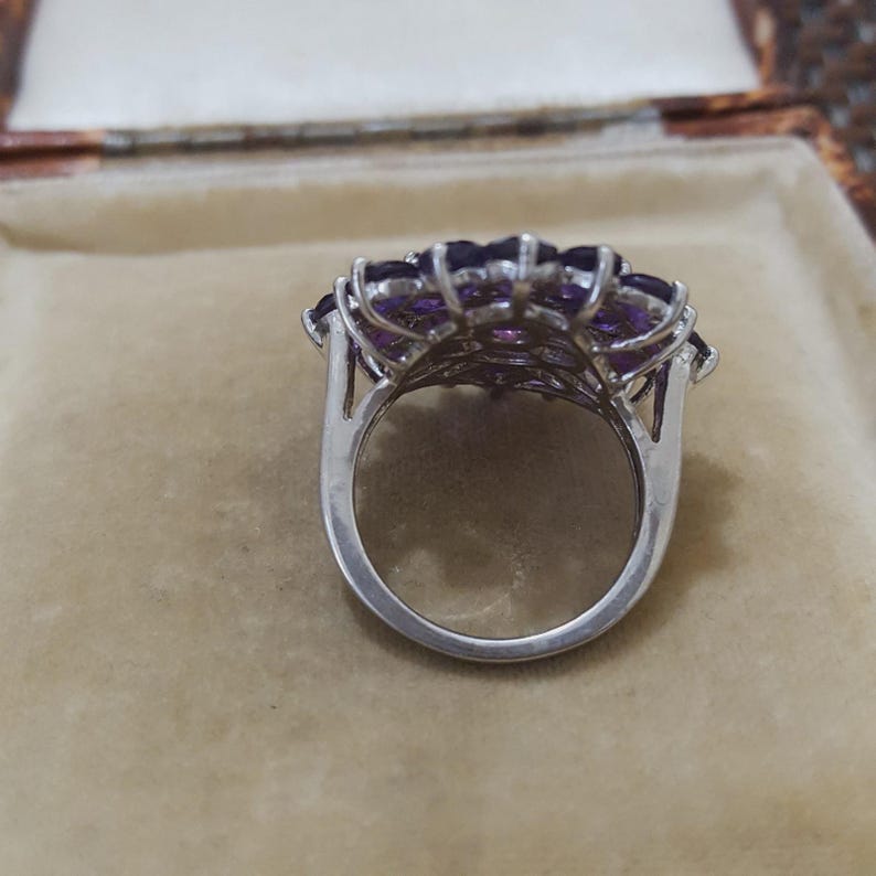 K&ouml;nnte beinhalten: Ein silberner Ring mit einem Cluster aus lila Edelsteinen. Die Edelsteine sind in einem floralen Muster angeordnet, mit einem polierten Silberband. Der Ring ist vor einem neutralen Hintergrund platziert, der das Design und die Farbe des Rings hervorhebt.