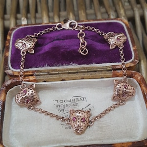 Può includere: Bracciale in oro rosa con sei ciondoli a forma di testa di leopardo con occhi di gemme rosa. Il bracciale è presentato in un portagioie vintage aperto con interno in velluto viola. La scatola è aperta.