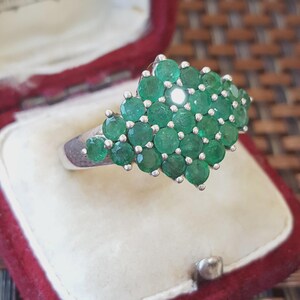 Puede incluir: Un anillo de plata con un grupo de numerosas piedras preciosas redondas de color verde vibrante dispuestas en una formación densa y multicapa. Las piedras están engastadas en una banda metálica, creando una superficie texturizada prominente.