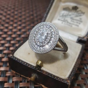 Op de afbeelding: Een zilveren ring met een grote, ronde kop, bezet met kleine, heldere edelstenen. Het midden heeft een grotere, ovaalvormige steen. De ring wordt gepresenteerd in een vintage, bruine sieradendoos.