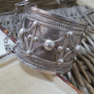 May include: A wide, silver cuff bracelet with an ornate design. The bracelet features raised circular and teardrop-shaped embellishments, connected by curved lines. A delicate chain is attached to the bracelet. The metal has a slightly aged appearance.