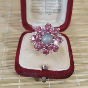 May include: A silver ring featuring a central grey gemstone, encircled by pink gemstones in a flower-like pattern. The ring is presented in a red velvet jewellery box, offering a vivid contrast. The ring's design is detailed and visually appealing.