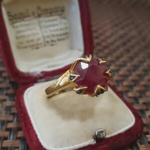 May include: A gold ring with a large, faceted ruby gemstone. The ruby is a deep red color and is surrounded by smaller, dark gemstones. The ring is displayed in a red velvet ring box.