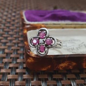 Pode incluir: Anel de prata com design de trevo de quatro folhas, com pedras preciosas de rubi ovais e um pequeno diamante. O anel é apresentado numa caixa de joias vintage com interior roxo.