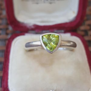 以下が含まれることがあります： 三角形にカットされた明るい緑色の宝石が滑らかな金属製のベゼルにセットされたシルバーリング。クリーム色の内装と深い赤色のベルベットの縁取りが施された小さな開いたジュエリーボックスの中にリングが収められています。