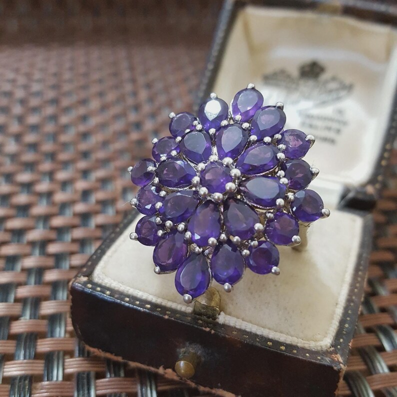 K&ouml;nnte beinhalten: Ein Silberring mit einem Cluster aus tropfenf&ouml;rmigen, tiefvioletten Edelsteinen. Der Ring ist in einem floralen Muster gefasst, wobei jeder Stein sorgf&auml;ltig platziert ist. Der Ring wird in einer Vintage-Schmuckschatulle in Braun pr&auml;sentiert.