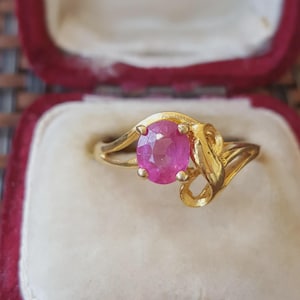 May include: A gold ring featuring a central oval-cut pink gemstone held by four prongs. The band has a polished gold finish and a swirling, asymmetrical design that curves around the stone, creating an open, flowing look on both sides.