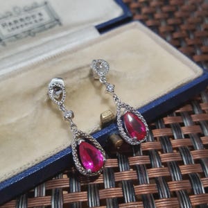 Könnte beinhalten: Ein Paar silberfarbene Ohrhänger mit einem tropfenförmigen, leuchtend rosa Edelstein. Die Edelsteine sind von einem Kranz aus kleinen, klaren Steinen umgeben. Die Ohrringe werden in einer blauen Schmuckschatulle präsentiert.