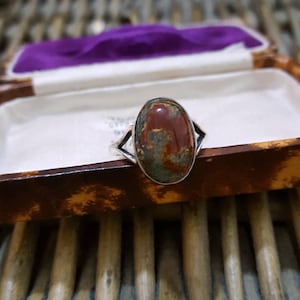 May include: A silver ring with an oval gemstone in shades of red, brown, and green. The ring has a silver band with a decorative design on either side of the stone. The ring is presented in a vintage jewelry box with a purple interior.