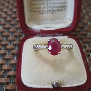 Puede incluir: Un anillo de plata con una gran gema de rubí ovalada, engastada en una banda retorcida. El anillo se exhibe en un joyero de terciopelo rojo con un interior color crema. El rubí es de color rojo intenso.