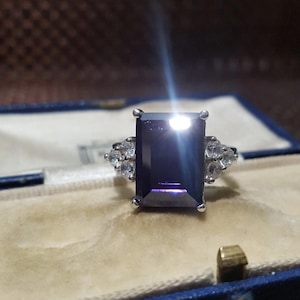 Puede incluir: Un anillo de plata con una gran piedra rectangular de color púrpura oscuro como pieza central. El anillo está flanqueado por piedras preciosas más pequeñas y claras a cada lado. El anillo se muestra en una caja de joyería azul con interior crema.
