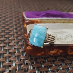 May include: A silver ring with a large, rectangular, light blue gemstone. The ring has a ribbed band and is set in a four-prong setting. The gemstone has a textured surface and a light blue color.