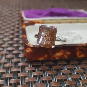 Pode incluir: Um anel de prata com uma grande pedra preciosa retangular marrom. A pedra preciosa tem uma rede de inclusões douradas. O anel está em uma caixa de joias vintage com interior roxo.