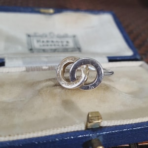 May include: A silver ring with two interlocking circles, displayed in a vintage blue jewellery box with a light blue velvet lining. The ring has a polished finish, showcasing a classic and elegant design. The ring is a timeless piece.