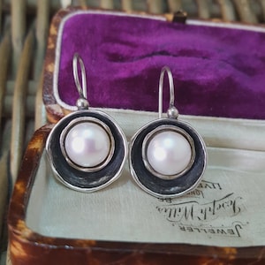 Op de afbeelding: Zilveren oorhangers met een centrale witte parel omgeven door een donkergrijs cirkelvormig frame. De oorbellen worden gepresenteerd in een vintage sieradendoosje met een paarse fluwelen binnenkant.