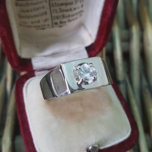 May include: A silver ring with a square face, featuring a central, round, clear gemstone. The ring is presented in a red velvet jewellery box with a cream interior. The ring's band is thick and polished.
