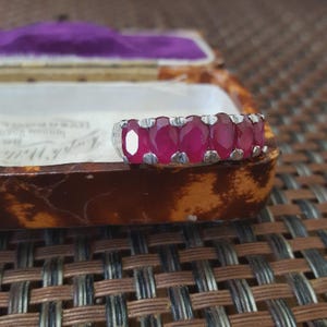 Op de afbeelding: Een zilveren ring met zes ovale, levendige rode edelstenen. De ring wordt gepresenteerd tegen een achtergrond van een vintage sieradendoosje met een paars interieur, wat het ontwerp en de kleur van de ring benadrukt.