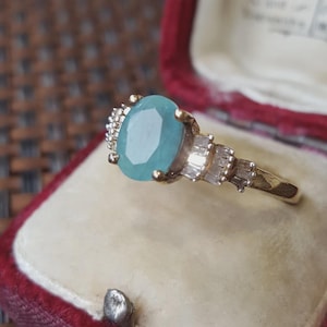 May include: A gold ring featuring a central oval-cut light blue gemstone held by four prongs, flanked by a series of small, clear, rectangular baguette-cut stones set along the band on both sides of the main setting.