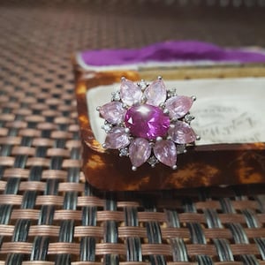 Op de afbeelding: Een zilveren ring met een bloemmotief. De centrale steen is een grote, ovale, dieproze edelsteen, omringd door kleinere, bloemblad-vormige roze en lichtroze stenen. Kleine heldere stenen zijn tussen de bloemblaadjes gezet. De ring wordt gepresenteerd in een vintage sieradendoosje.