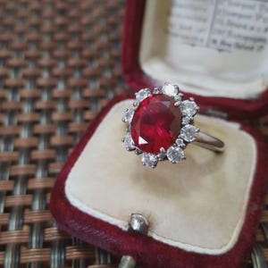 Op de afbeelding: Een zilveren ring met een grote, ovale, rode edelsteen, omringd door kleinere, ronde, heldere edelstenen. De ring wordt gepresenteerd in een rode fluwelen ringdoos met een crèmekleurig interieur. Het ontwerp van de ring is klassiek en elegant.