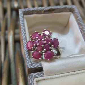 May include: A silver ring featuring a cluster of oval-cut, deep red rubies. The ring is set in a silver band and is displayed inside a vintage gray jewelry box with a white satin lining. The rubies are the focal point.