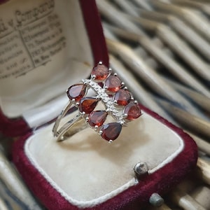 Può includere: Un anello d'argento con un gruppo di pietre preziose rosse a forma di goccia. L'anello presenta una doppia fascia e un design centrale delle pietre preziose disposte a forma quadrata, con pietre chiare più piccole tra loro. L'anello è esposto in un portagioie di velluto bordeaux.
