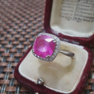 Puede incluir: Un anillo plateado con una gran piedra preciosa rosa brillante de forma cuadrada. La piedra preciosa está rodeada por un halo de piedras más pequeñas y transparentes. El anillo se exhibe en un joyero de terciopelo burdeos.