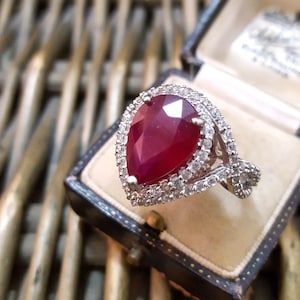 Op de afbeelding: Een ring met een peervormige robijn, gezet in een zilveren band met een halo van kleinere diamanten. De ring bevindt zich in een zwarte fluwelen ringdoos.