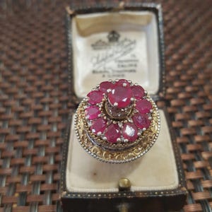 May include: A vintage ring with a cluster of oval-cut, deep red rubies set in a gold and silver filigree band. The ring is displayed in a small, antique jewelry box with a velvet interior.