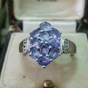 May include: A silver-toned ring featuring a cluster of nine oval-cut, light purple stones arranged in a diamond shape. The band is accented with small, clear, round stones on each side, set into the metal shank, creating a decorative finish.