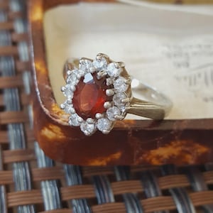 May include: A silver-toned ring featuring a central oval-cut deep red gemstone surrounded by a halo of small, clear, round-cut stones. The ring has a polished metal band and is displayed in a decorative container with a light-colored interior.