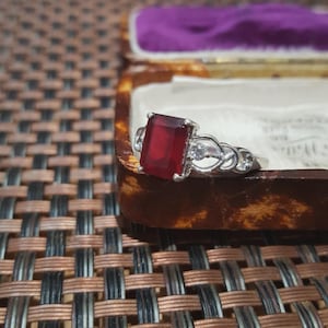 Könnte beinhalten: Ein silberner Ring mit einem großen, rechteckigen, tiefroten Edelstein. Ein kleiner, runder, klarer Stein ist daneben eingesetzt. Der Ring wird in einer Vintage-Schmuckschatulle mit lila Innenfutter präsentiert.