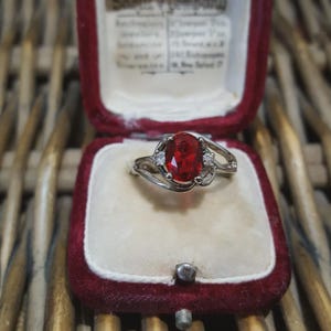Peut inclure: Une bague en argent ornée d'une grande pierre rouge ovale et de deux petites pierres claires. La bague est présentée dans un écrin à bijoux vintage en velours rouge. L'écrin est ouvert, révélant la bague sur l'intérieur en peluche.