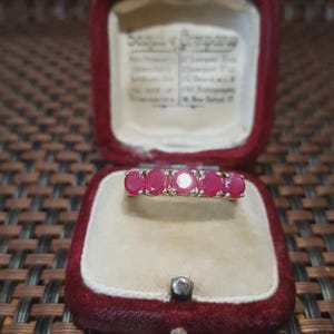 Peut inclure: Une bague en argent ornée de cinq pierres précieuses rouges vives, présentée dans un écrin à bijoux en velours rouge vintage. Les pierres sont espacées de manière uniforme, créant un design classique. L'intérieur de la boîte est crème.