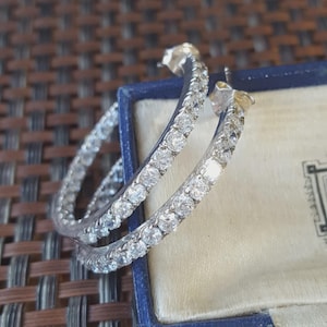 May include: A pair of silver-toned hoop earrings featuring a continuous row of clear, round-cut stones set along the outer edge. The earrings are positioned partially inside a blue jewelry box with a cream-colored interior lining.