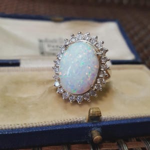 Könnte beinhalten: Ein goldener Ring mit einem großen, ovalen Opal-Edelstein, umgeben von kleinen, klaren Edelsteinen. Der Opal hat eine milchig-weiße Basis mit irisierenden Farbtönen. Der Ring ist in einem goldenen Band gefasst und wird in einer blauen Schmuckschatulle präsentiert.