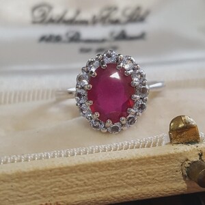 Pode incluir: Um anel de tom prateado com uma grande pedra preciosa de cor vermelho-escura e corte oval no centro, rodeada por um halo de pequenas pedras claras de corte redondo. O anel está sobre o interior de tecido creme de uma caixa de joias.