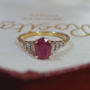 May include: A gold ring featuring a central oval-cut red ruby gemstone held by four prongs. The band is accented on each side by a cluster of three small, clear baguette-cut diamonds set in a row, creating a symmetrical and refined appearance.